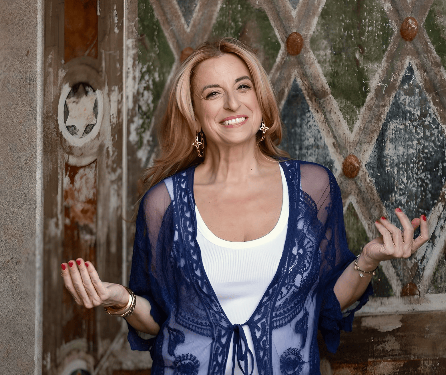 Nathalie Botros, the Bon-Vivant Girl and Happiness Activator, radiates confidence and warmth as she smiles brightly. Dressed in a stylish navy blue lace cover-up over a white outfit, she strikes a relaxed pose against an ornate, vintage-style backdrop. Her engaging expression and open gesture reflect her role as an author, psychotherapist, motivational speaker, and mindset coach, inspiring others to embrace happiness and live authentically.